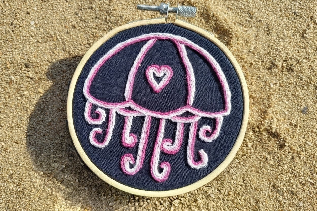 Embroidery hoop with a pink jellyfish design on a dark background, placed on sand.